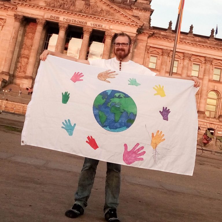 Planetarische weiße Flagge © Alexander Donath Mann hält eine Flagge mit bunten Handabdrücken und der Erde vor dem Reichstag.